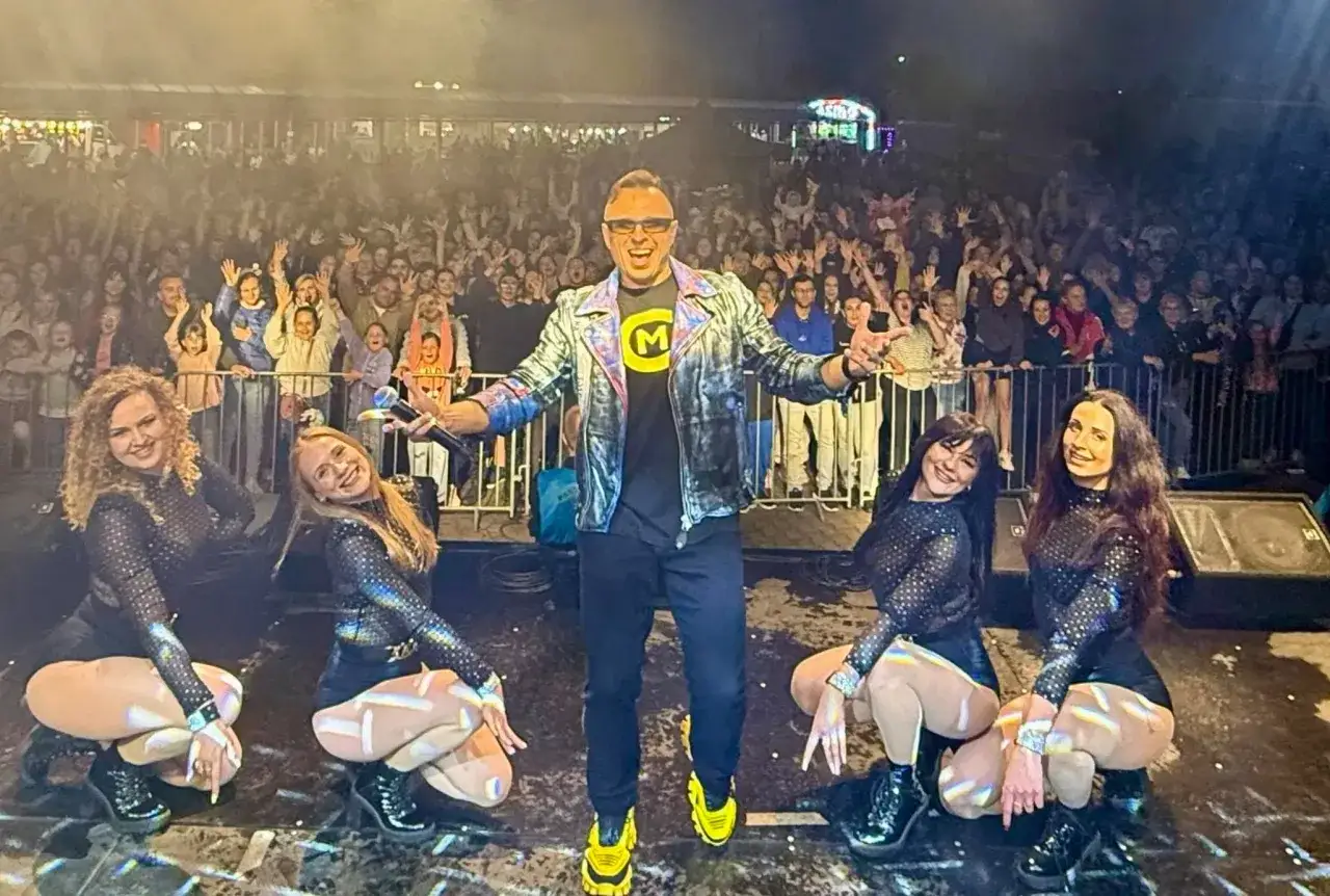 Gwiazdor disco polo w błyszczącej kurtce i tancerki w błyszczących strojach na scenie, przed roztańczonym tłumem. Imperium disco polo w pełnej krasie!