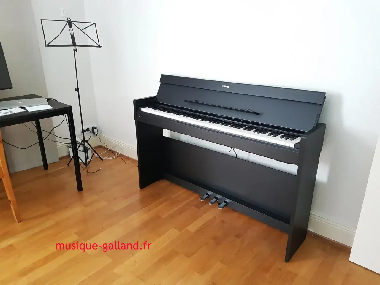 Un piano num&eacute;rique Yamaha noir, pr&ecirc;t &agrave; jouer, avec un pupitre et un bureau &agrave; proximit&eacute;.