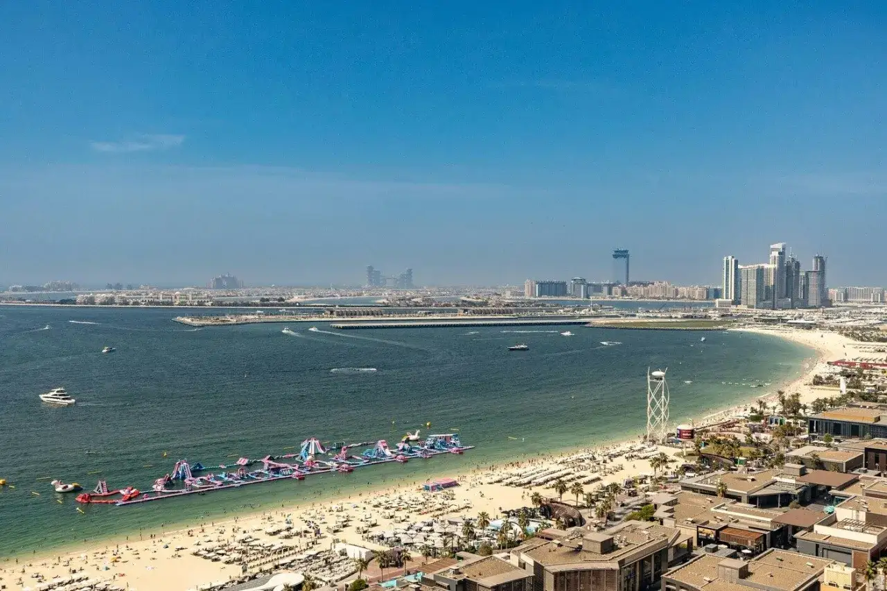 Wakacje w Dubaju: plaża z dmuchanymi atrakcjami wodnymi, tłumy ludzi, łodzie i nowoczesne wieżowce w tle.