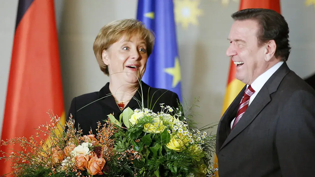 Angela Merkel, die erste Bundeskanzlerin Deutschlands, lächelt mit einem Blumenstrauß. Neben ihr steht ein lachender Mann.