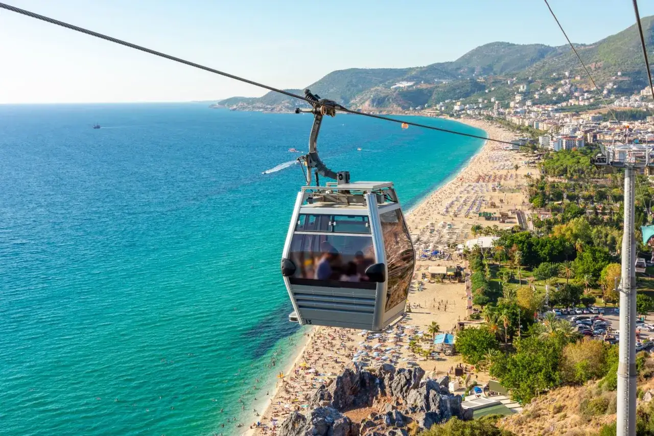 Seilbahn über dem belebten Alanya Strand mit türkisfarbenem Meer und vielen Sonnenanbetern.