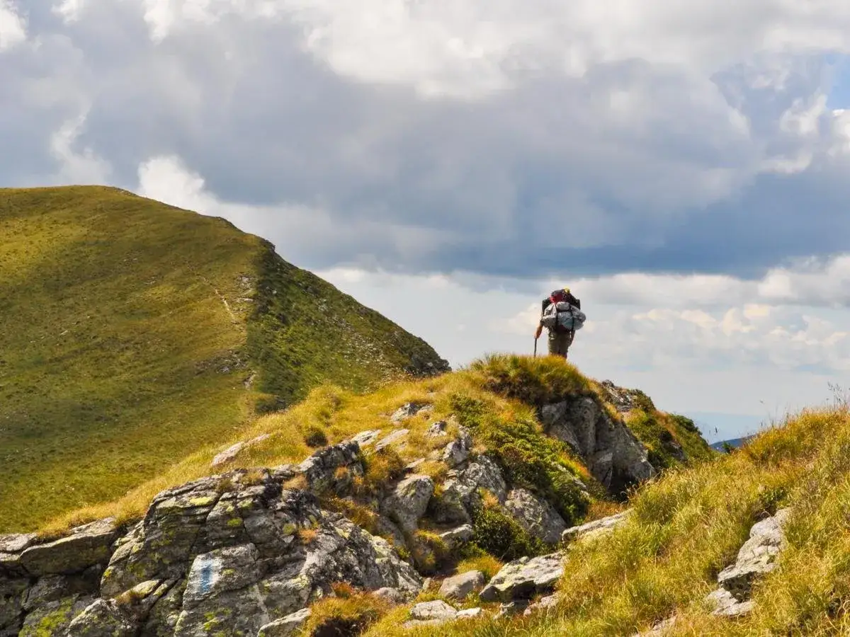 Ein Wanderer mit Rucksack und Stock steht auf einem felsigen Grat. Im Hintergrund erstreckt sich ein grüner Berghang. Ein tolles Erlebnis beim **Karpaten wandern**.