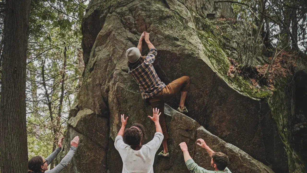 En person klättrar på en stor sten vid Kjugekull, medan vänner ger stöd. Bouldering i naturen.