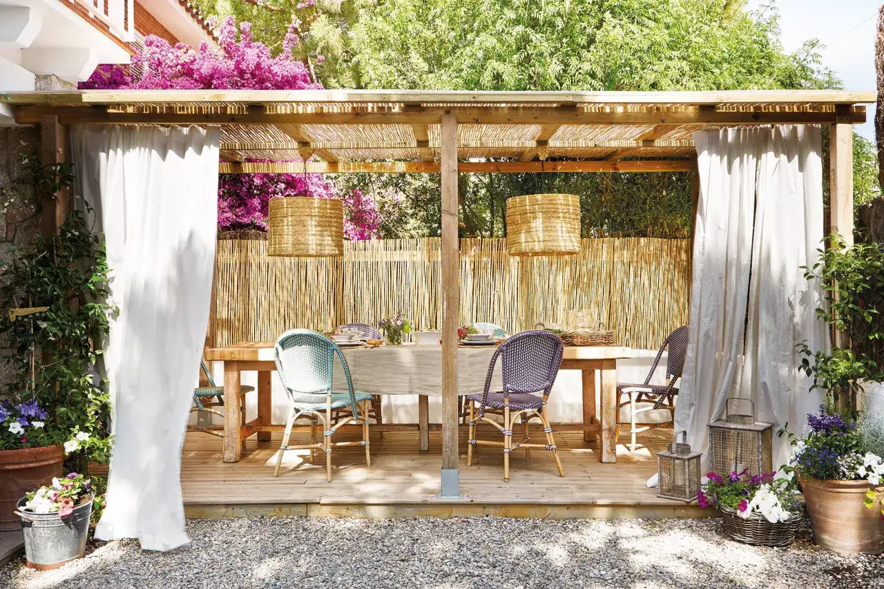 Pérgola de madera con enredaderas para pergolas, cortinas blancas y mesa puesta para comer al aire libre.