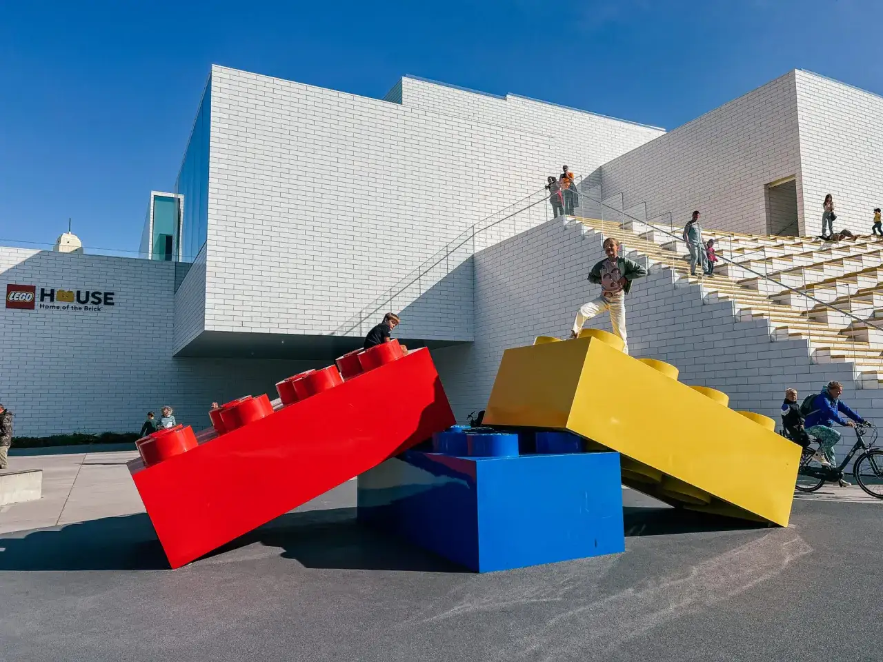 Gigantyczne klocki Lego przed budynkiem Lego House, gdzie można kupić bilety. Ludzie spacerują po schodach i bawią się na zewnątrz.