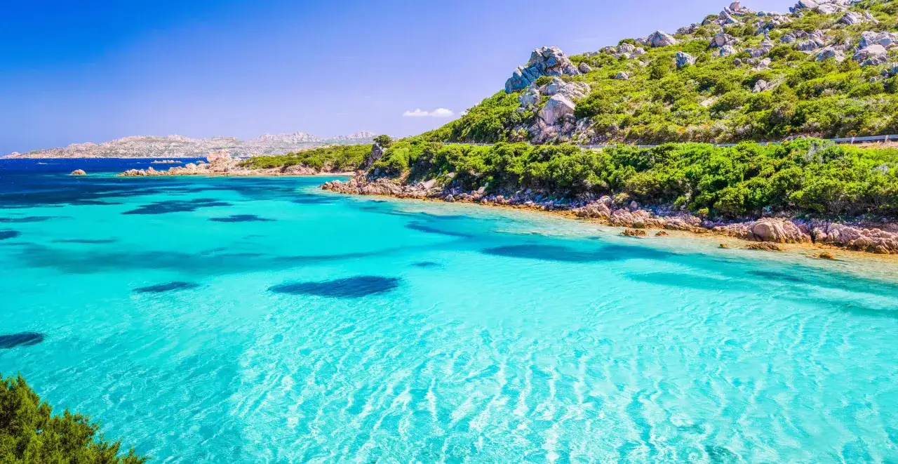 Kristallklart turkost vatten vid en gr&ouml;nskande kustlinje med klippor. Perfekt f&ouml;r en resa till Sardinien, h&auml;r &auml;r n&aring;gra tips f&ouml;r din semester.
