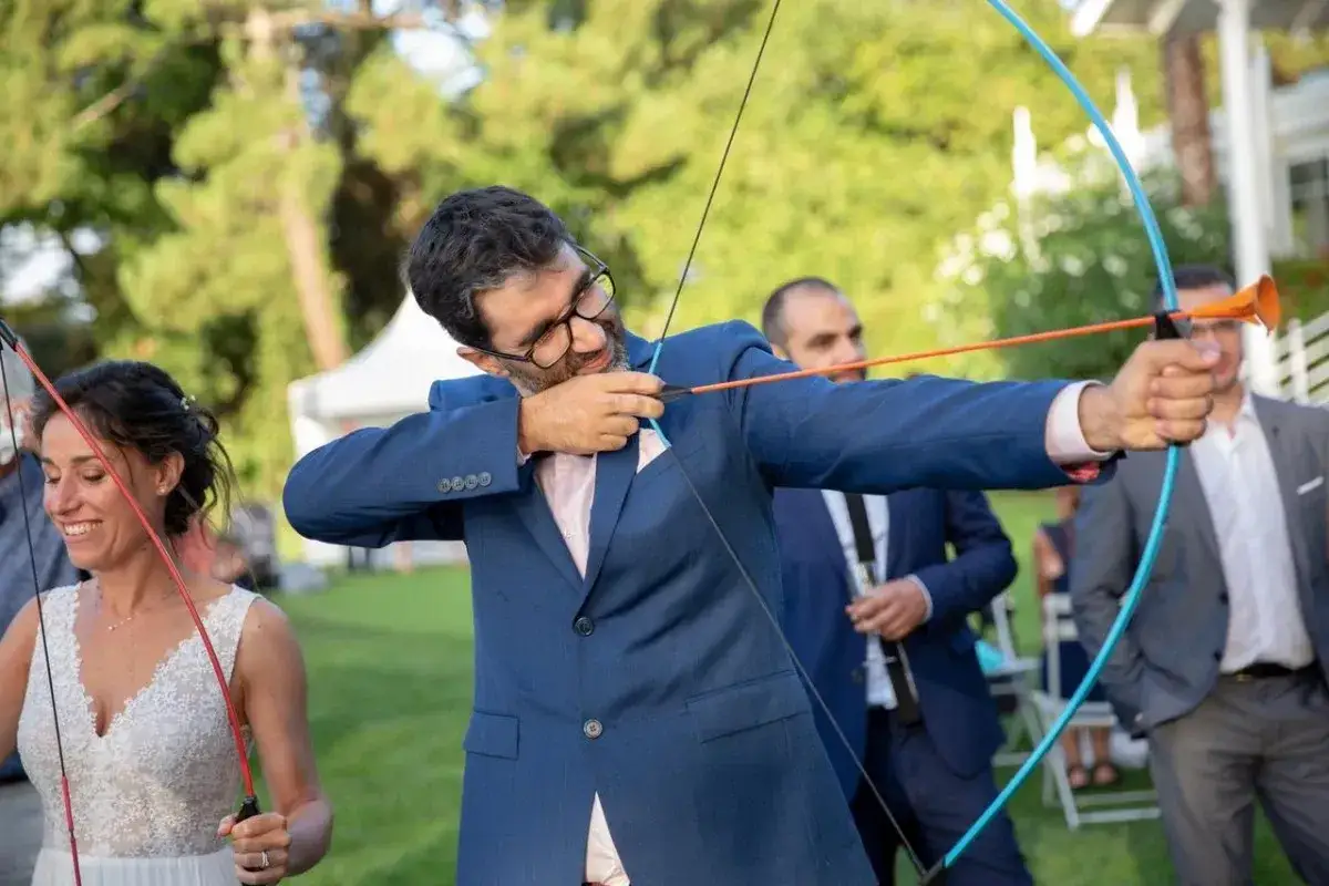 Novio con arco y flecha, parte de los juegos de bodas. La novia sonr&iacute;e al fondo.