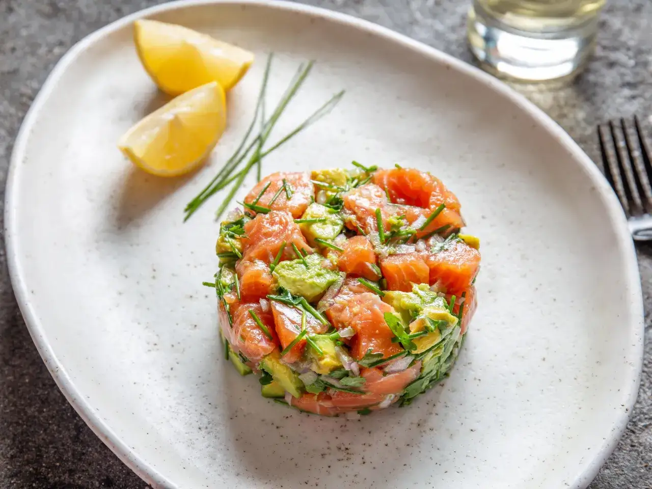 Un d&eacute;licieux tartare de saumon frais, m&eacute;lang&eacute; &agrave; de l'avocat et des herbes, servi avec des quartiers de citron sur une assiette blanche.