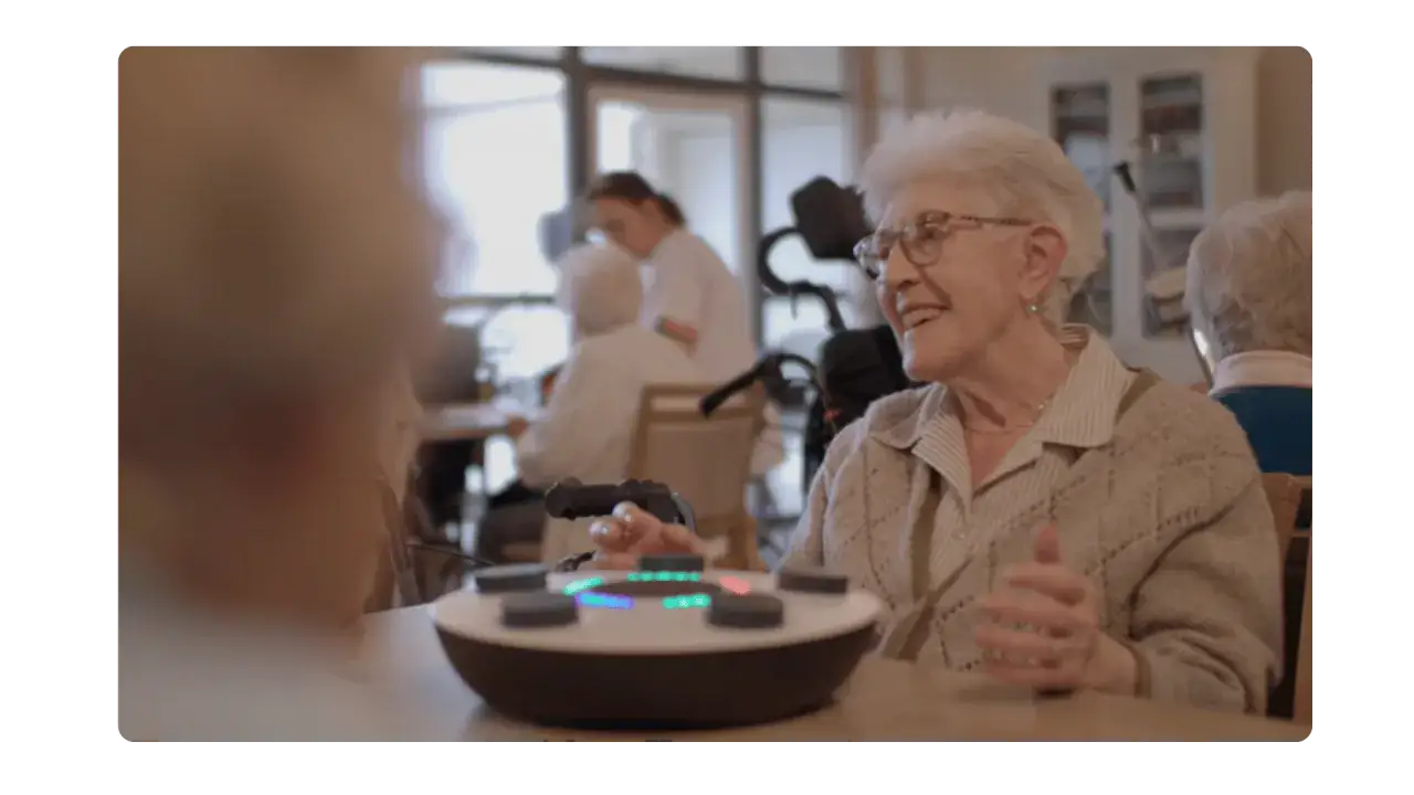 Une dame &acirc;g&eacute;e souriante participe &agrave; un atelier sensoriel. Elle interagit avec un appareil interactif lumineux, stimulant ses sens.