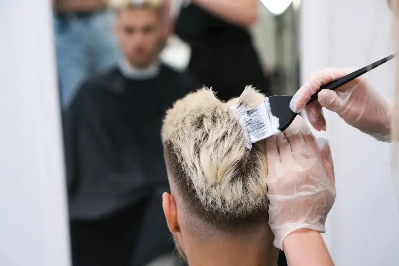 Fryzjerka w rękawiczkach nakłada farbę na blond włosy mężczyzny. Nowoczesne farbowanie włosów męskich w salonie.