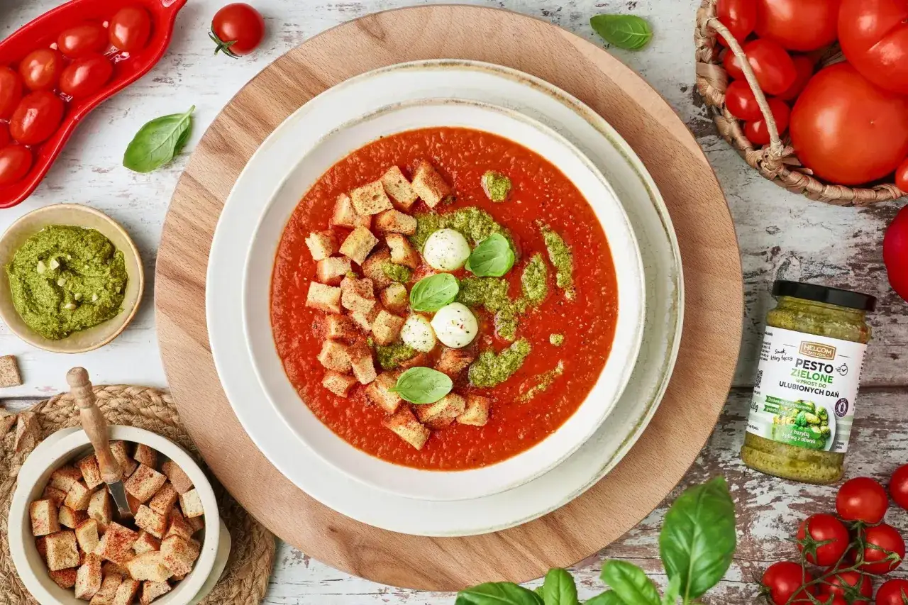 Zupa pomidorowa z grzankami, kulkami mozzarelli i zielonym pesto. Obok słoiczek pesto pomidorowego.