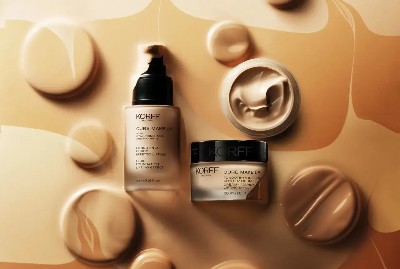 Due flaconi di base trucco KORFF, uno fluido e uno in crema, immersi in una texture cremosa e dorata.
