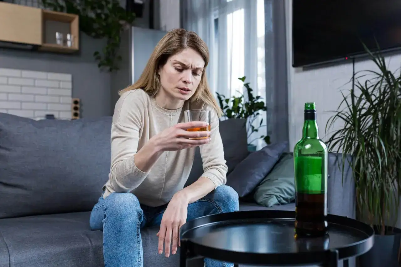 Kobieta siedzi sama na kanapie, trzymając szklankę z bursztynowym płynem. Obok stoi otwarta butelka. To smutne picie w samotności.