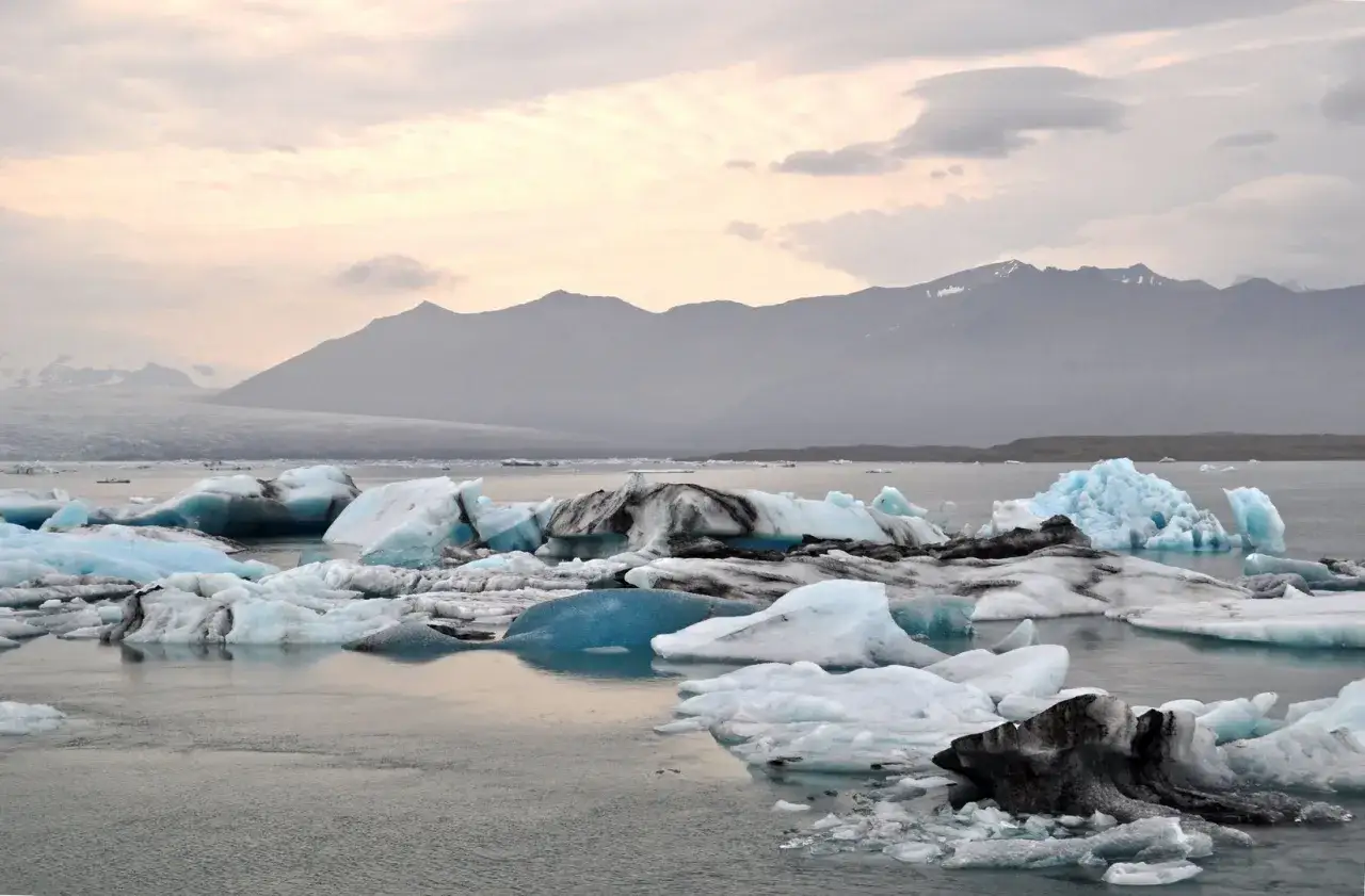 Islandia lodowce: góra lodowa o turkusowym odcieniu i inne fragmenty lodowców unoszą się na spokojnej wodzie, w tle mgliste góry.