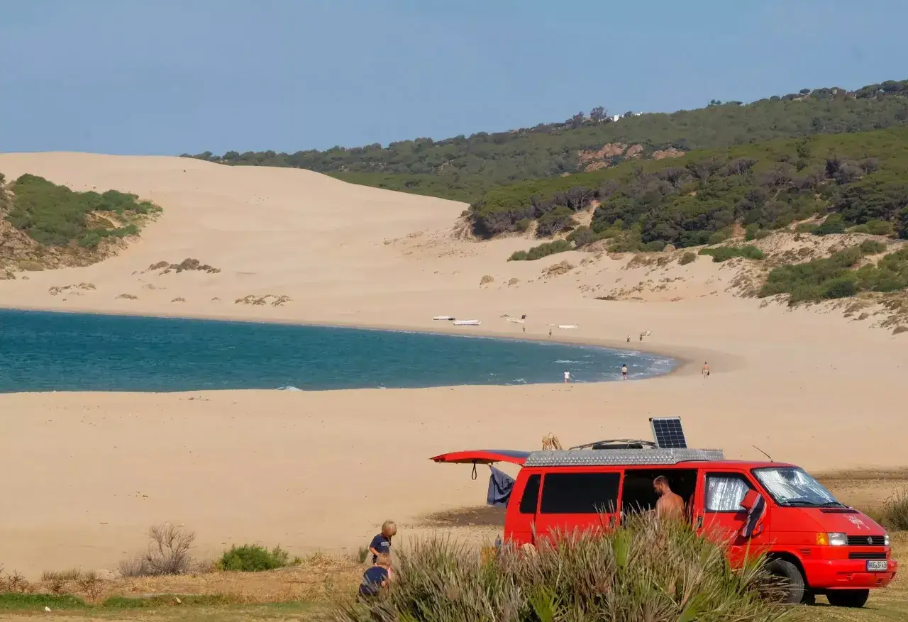 Una furgoneta roja aparcada en la playa del &aacute;rea Bolonia, con dunas y mar azul al fondo.
