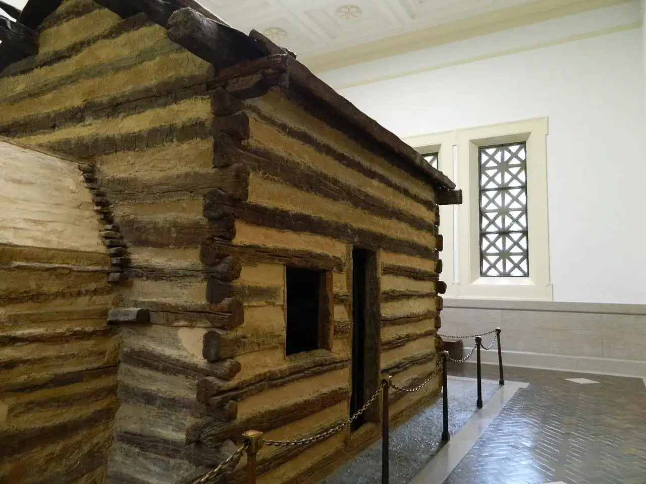 Maquette d'une cabane en rondins, &eacute;voquant l'abraham lincoln origine. Une fen&ecirc;tre d&eacute;corative et un sol carrel&eacute; compl&egrave;tent la sc&egrave;ne.
