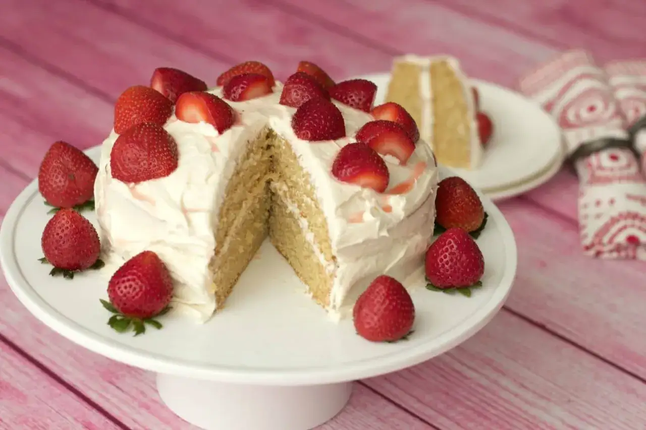 Ein k&ouml;stlicher veganer Kuchen mit wei&szlig;em Frosting und frischen Erdbeeren. Ein St&uuml;ck ist bereits angeschnitten.
