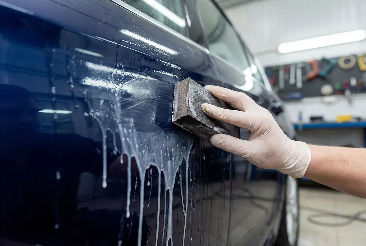 Gloved hand uses a sanding block to restore clear coat on a dark blue car, creating a foamy lather.