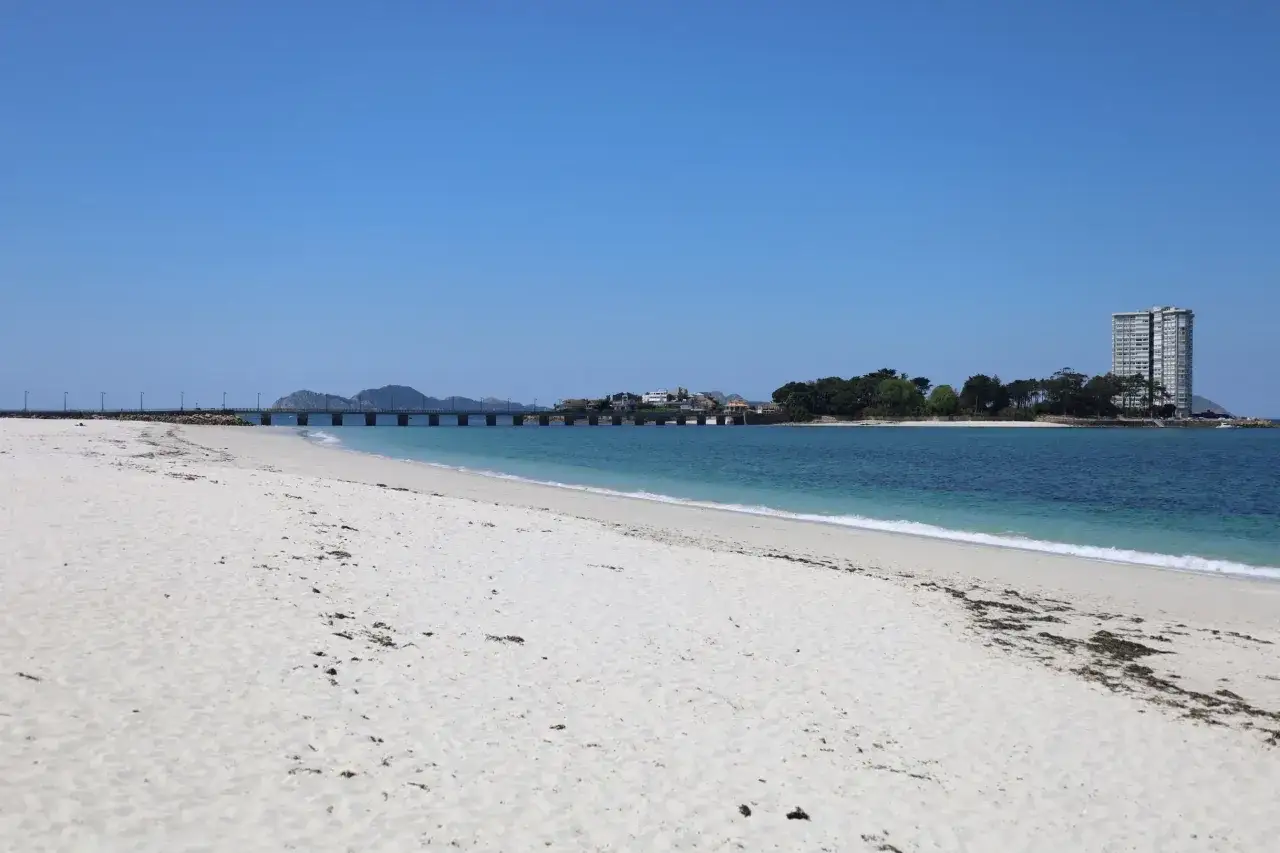 Amplia playa de arena blanca en la playa del Vao, Vigo, con el mar azul y un puente al fondo.