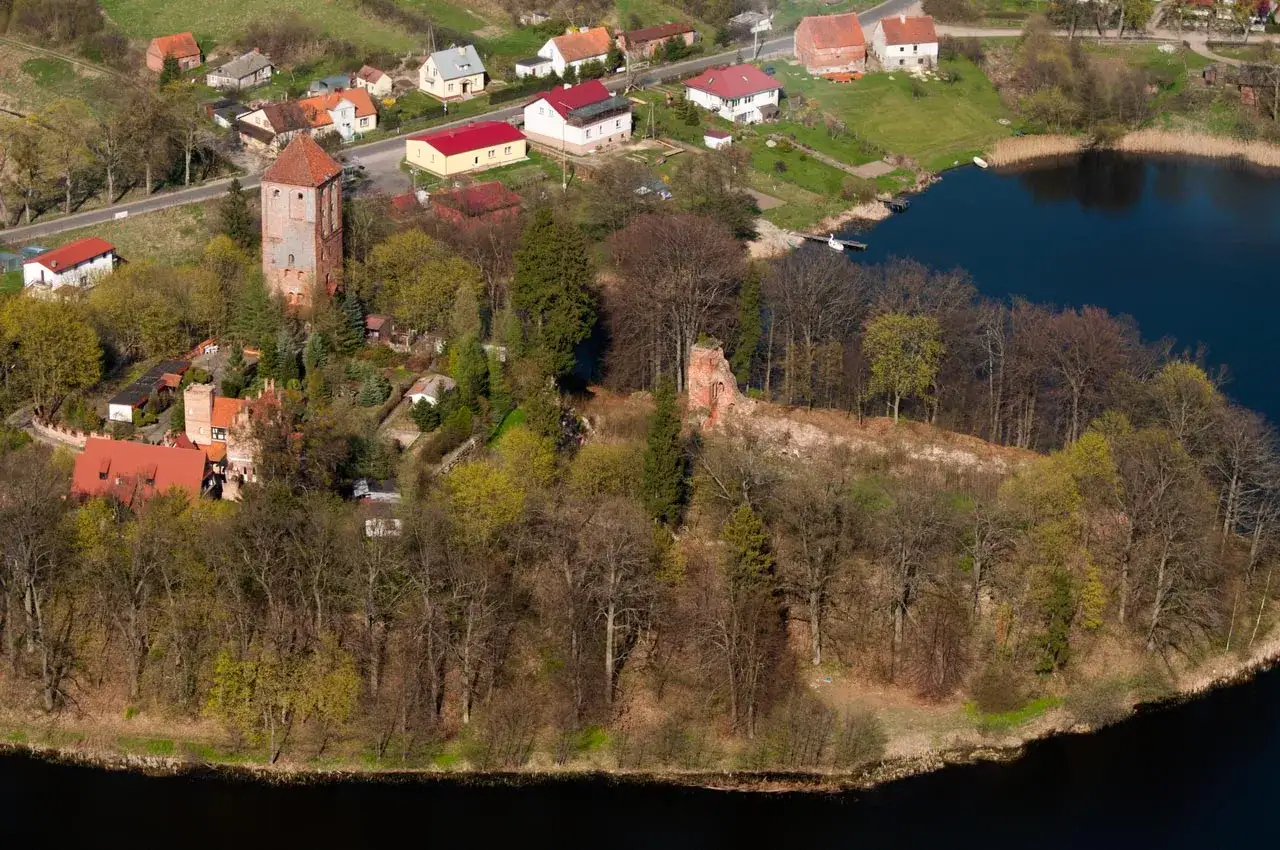 Przezmark zamek, wieża i ruiny na wzg&oacute;rzu nad jeziorem, otoczone drzewami i zabudowaniami.
