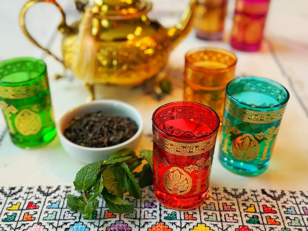 Service à thé marocain : thé vert, menthe fraîche, verres colorés et théière dorée sur une nappe brodée.