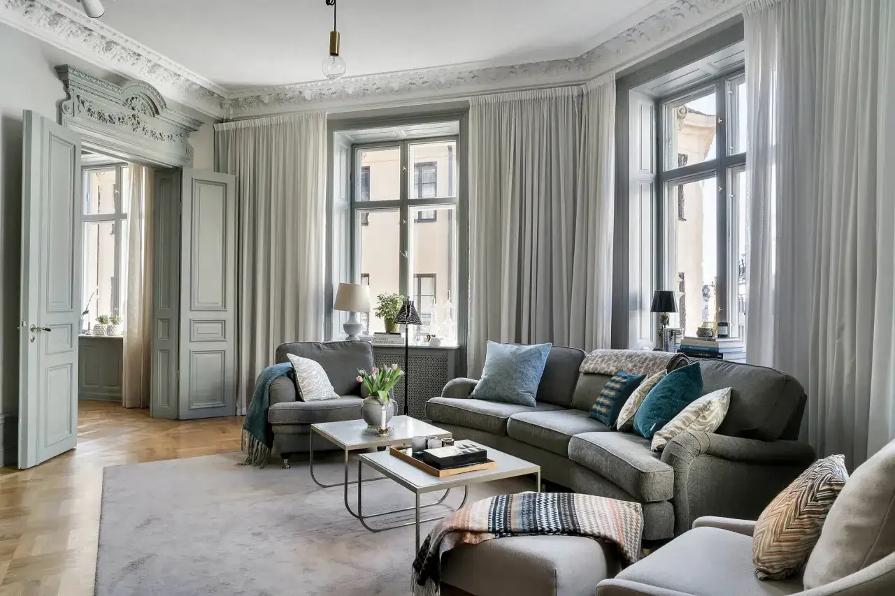 Sal&oacute;n elegante con sof&aacute;s grises, mesas de centro y alfombra. Las puertas dobles y las ventanas altas permiten distribuir salones irregulares, creando un ambiente acogedor.