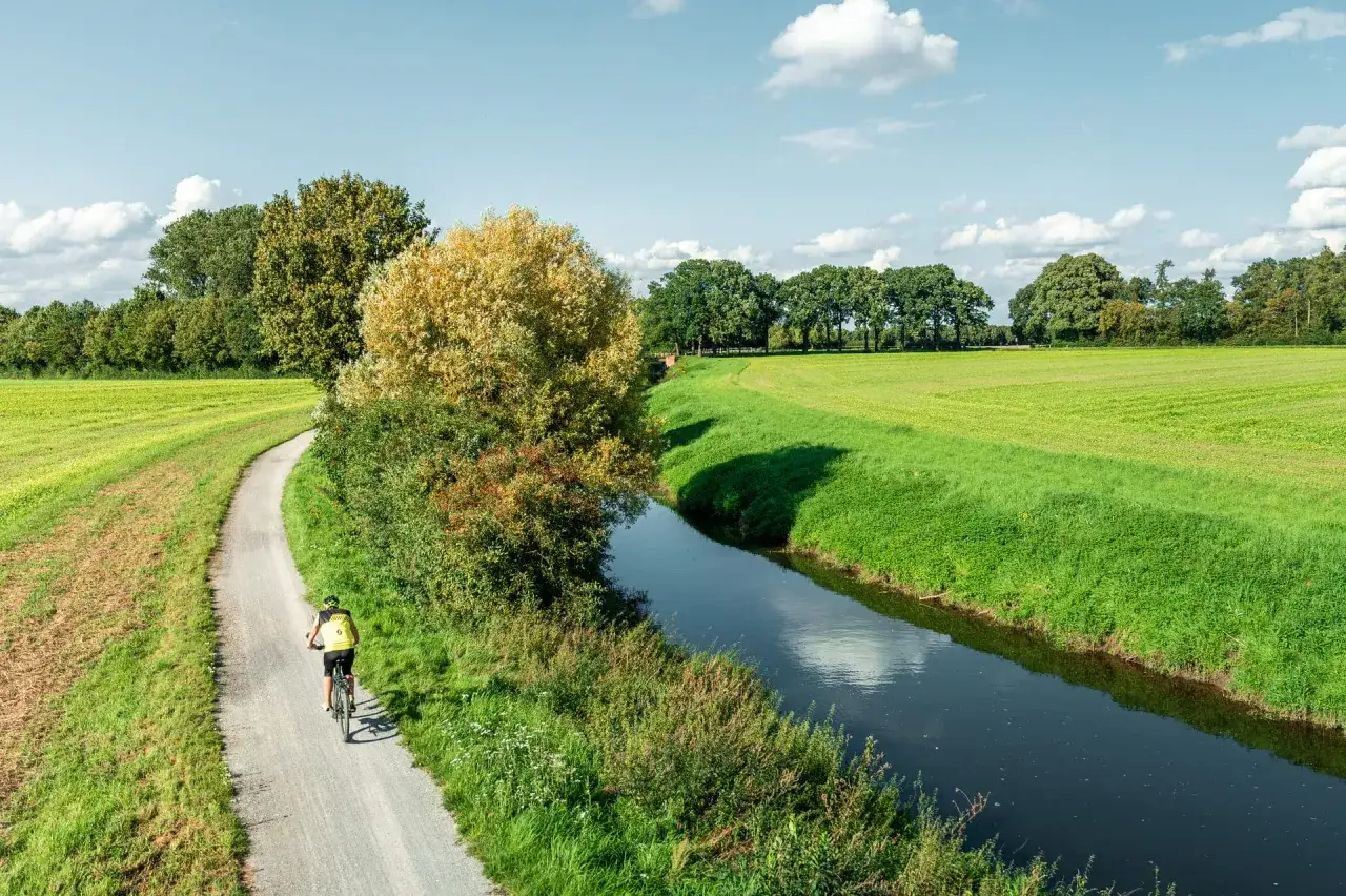 Radfahrer auf dem werse ems radweg, vorbei an gr&uuml;nen Feldern und einem Fluss unter blauem Himmel mit Wolken.