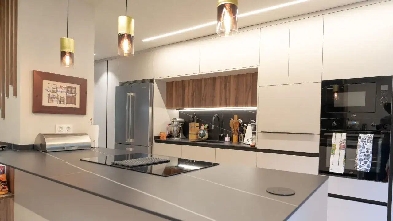 Cocina moderna con isla central. La iluminaci&oacute;n c&aacute;lida de las l&aacute;mparas colgantes y la luz lineal bajo los gabinetes demuestran que luz es mejor para la cocina.
