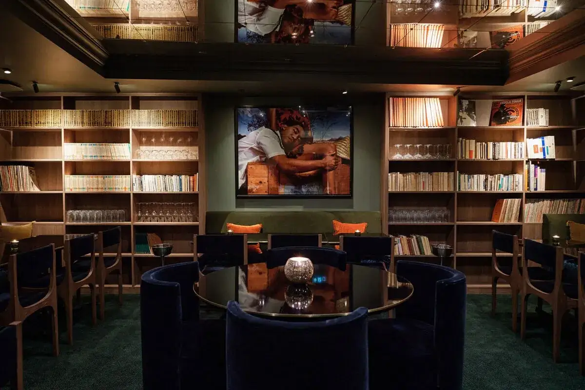 Un bar cosy, ambiance feutrée synonyme, avec des étagères remplies de livres et de verres, une grande peinture au centre, et des fauteuils en velours bleu nuit.