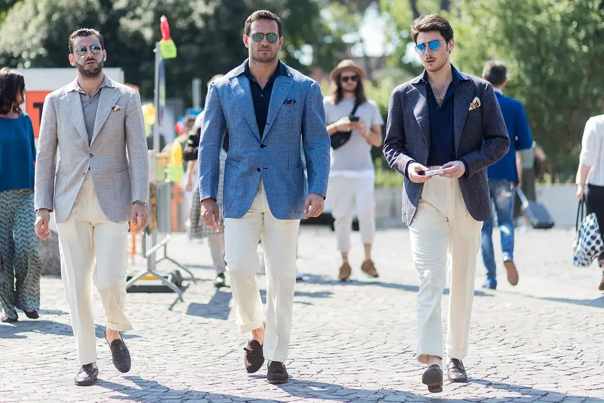 Drei Männer in eleganten Outfits: Ein Mann trägt ein blaues Sakko mit beige Hose, ein anderer ein helles Sakko mit beige Hose.