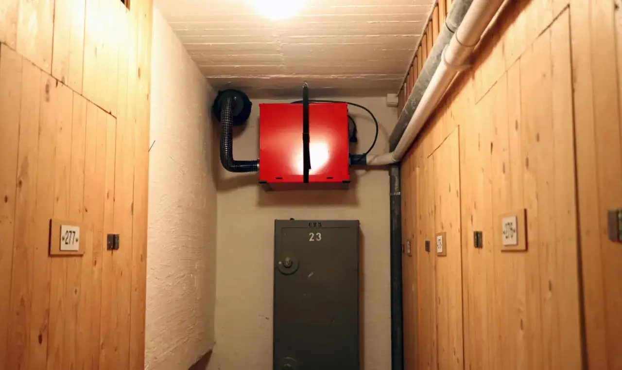 Röd radonfläkt monterad på väggen i en källarkorridor med träpaneler och numrerade dörrar.