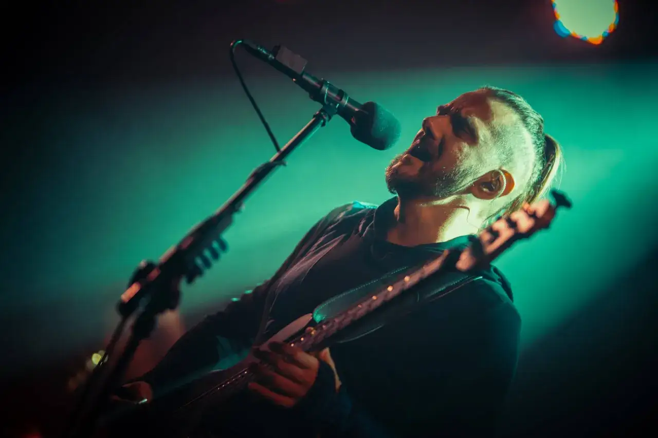 Wokalista z dredami śpiewa do mikrofonu, grając na gitarze. Jego emocje płyną jak rwąca denial river.