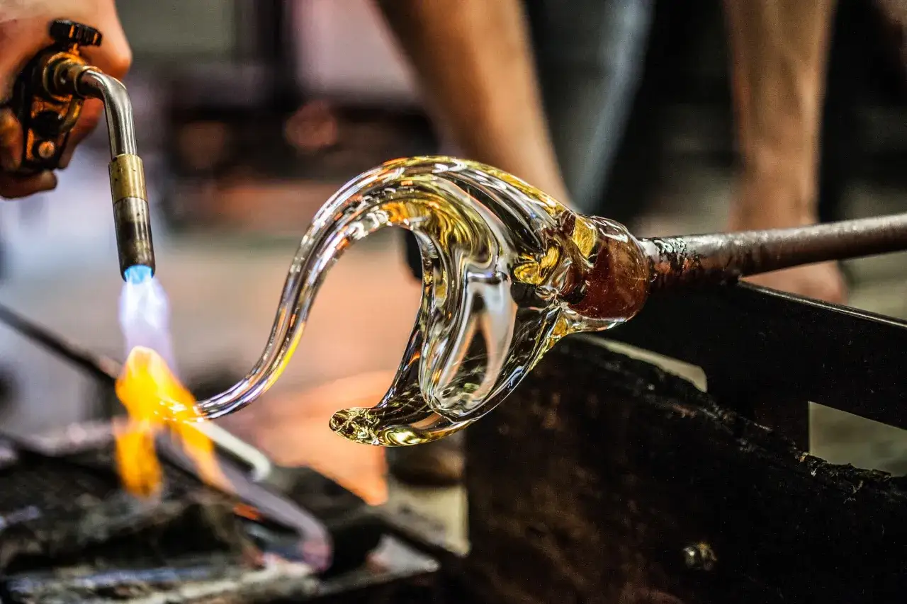 Un artisan verrier façonne le verre en fusion avec une torche. La flamme bleue et orange illumine la matière malléable, révélant son éclat.