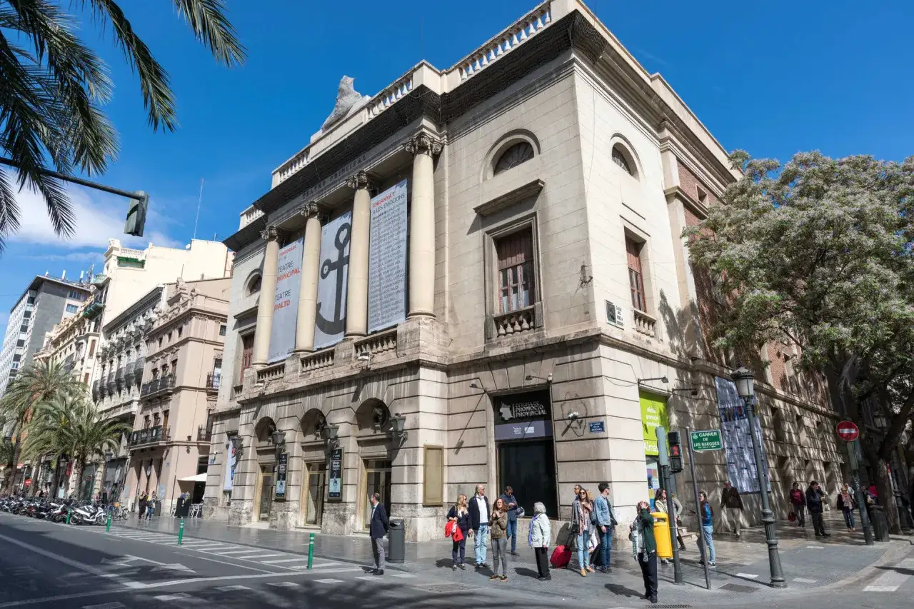 El **teatro en Valencia**, el Principal, con su fachada clásica y carteles anunciando espectáculos, se alza majestuoso bajo un cielo azul.