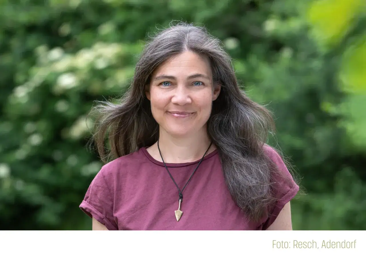 Martha Sophie Marcus lächelt in die Kamera, ihr langes Haar weht im Wind. Sie trägt ein weinrotes Shirt mit einem Pfeil-Anhänger.