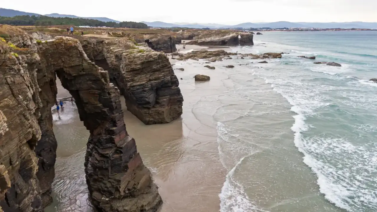 Arco natural en las playas de la Costa da Morte, con personas explorando la arena y el mar turquesa.