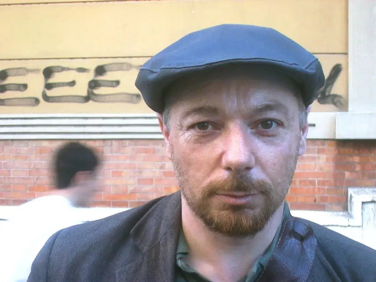 Uomo con berretto e barba, sullo sfondo graffiti e mattoni. Potrebbe essere Paolo Nori, autore di libri che invitano alla riflessione.