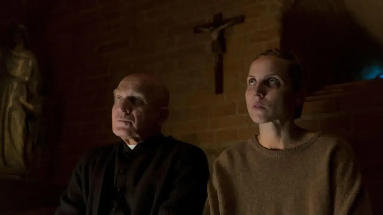 Sługi Boże film: starszy mężczyzna w sutannie i młoda kobieta patrzą w górę w mrocznym wnętrzu, nad nimi krzyż.