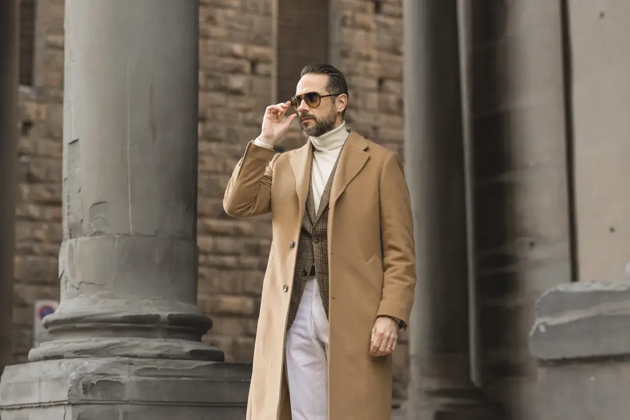 Mann im neuen Geld-Stil mit Kamelhaarmantel, Rollkragenpullover, karierter Weste und Sonnenbrille.