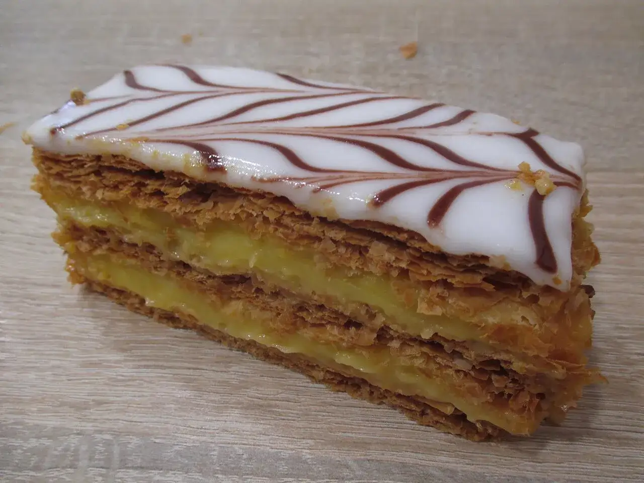 Une part de fondant millefeuille, avec ses feuilletages croustillants et sa crème pâtissière onctueuse, surmontée d'un glaçage blanc décoré.