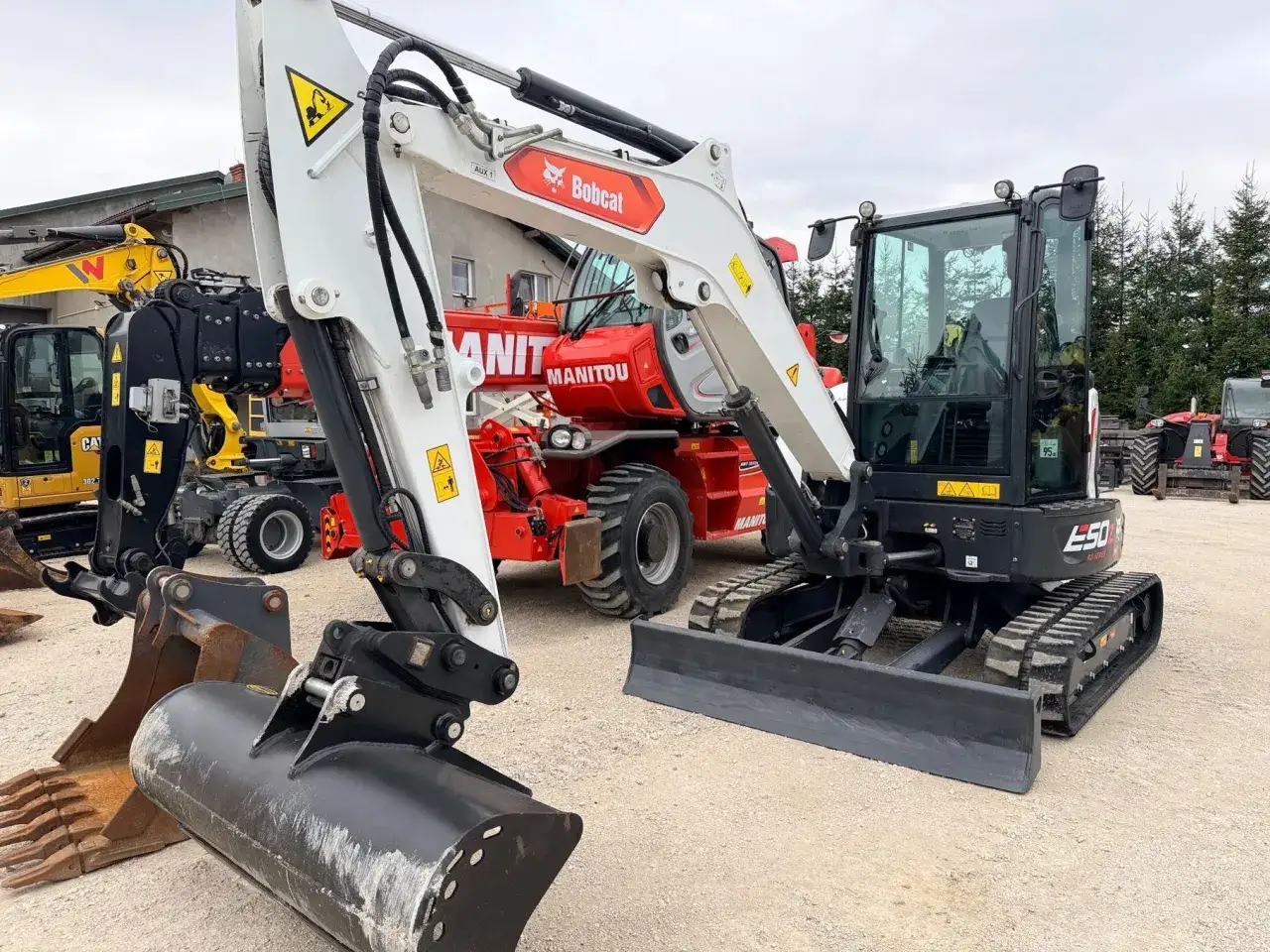 Biała mini koparka Bobcat E50 z czarnym lemieszem i gąsienicami. W tle czerwony ładowarka Manitou. Waga maszyny jest kluczowa przy transporcie.