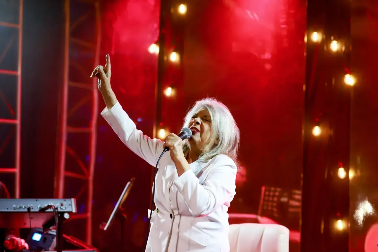 Krystyna Prońko, w białej marynarce, śpiewa na scenie. Jej ręka uniesiona w górę, jakby podkreślała emocje w jej utworach.
