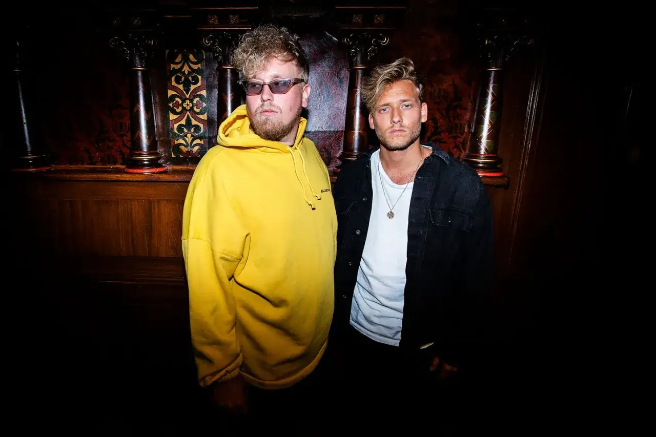 Sonny Fahlberg och en v&auml;n poserar i en m&ouml;rk milj&ouml; med tr&auml;paneler och utsmyckade pelare.
