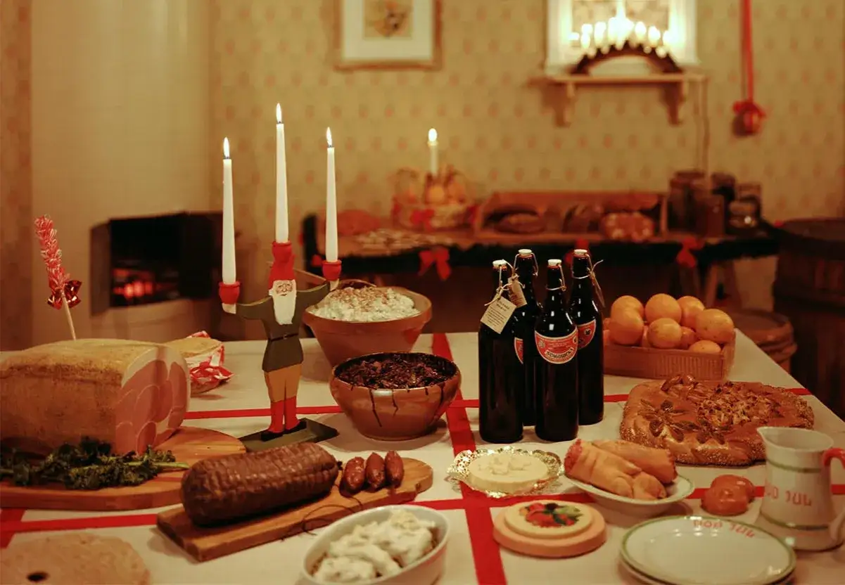 Ett dukat bord med julmat: skinka, korv, sill, risgrynsgröt, apelsiner, bröd och flaskor med dryck. En tomte med ljus står mitt på bordet. Här ser du vad som finns på julbordet.