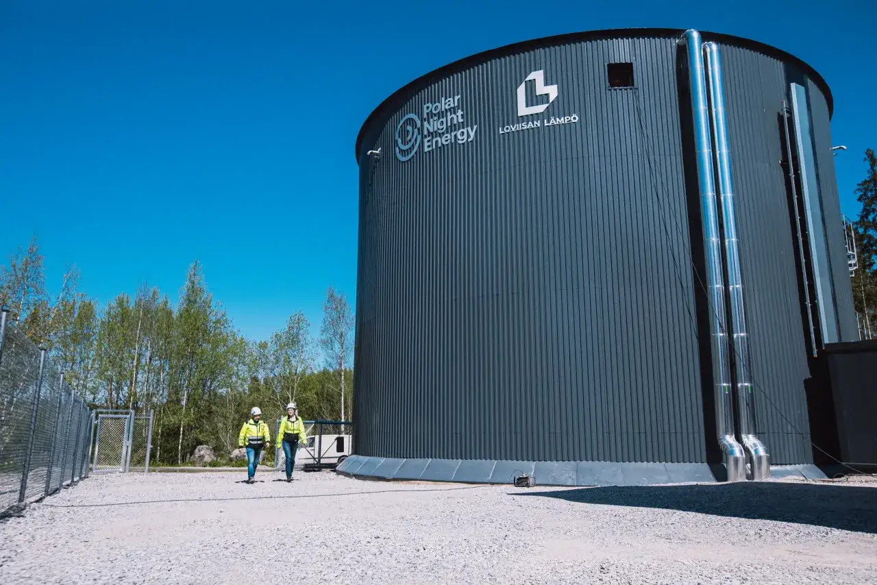 Wielki zbiornik Polar Night Energy, gdzie odbywa się magazynowanie ciepła w piasku. Dwóch pracowników w kamizelkach odblaskowych idzie obok.