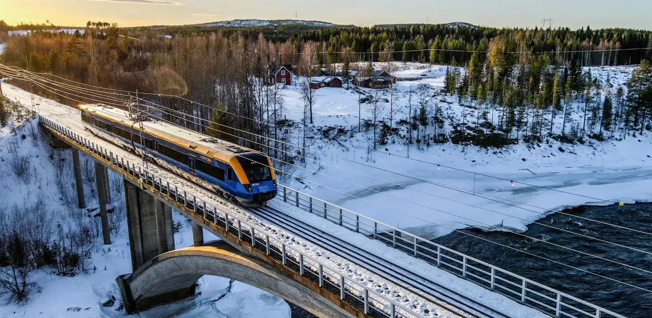 Blått och gult tåg på en bro över en frusen älv, på väg till Kalix genom ett snöigt vinterlandskap med skog och röda hus.