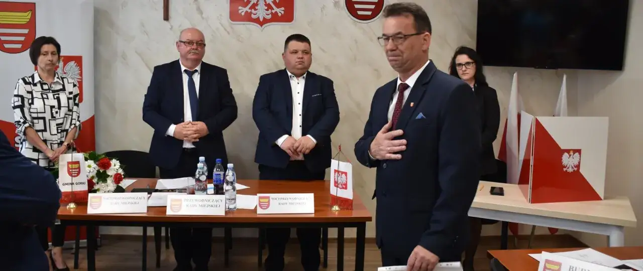 Przewodniczący rady miasta i jego zastępcy, w garniturach, stoją przy stole. Dyskutują o tym, ile zarabia przewodniczący rady miasta.