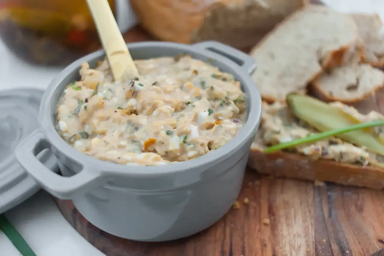 Pyszna pasta z tuńczyka i jajek w szarym naczyniu, obok kromki chleba z og&oacute;rkiem.