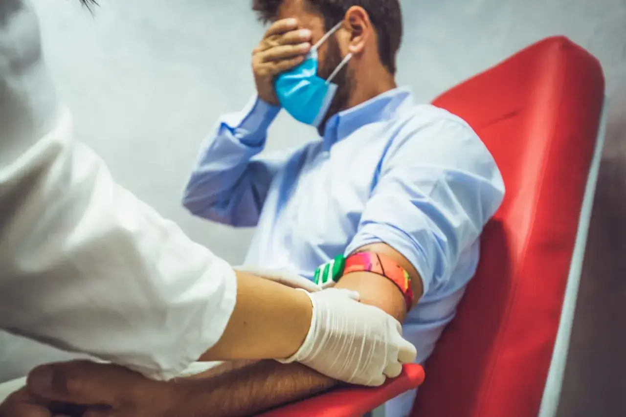 Man met fobie voor naalden bedekt ogen tijdens bloedafname. Een verpleegkundige met handschoenen bereidt de arm voor.