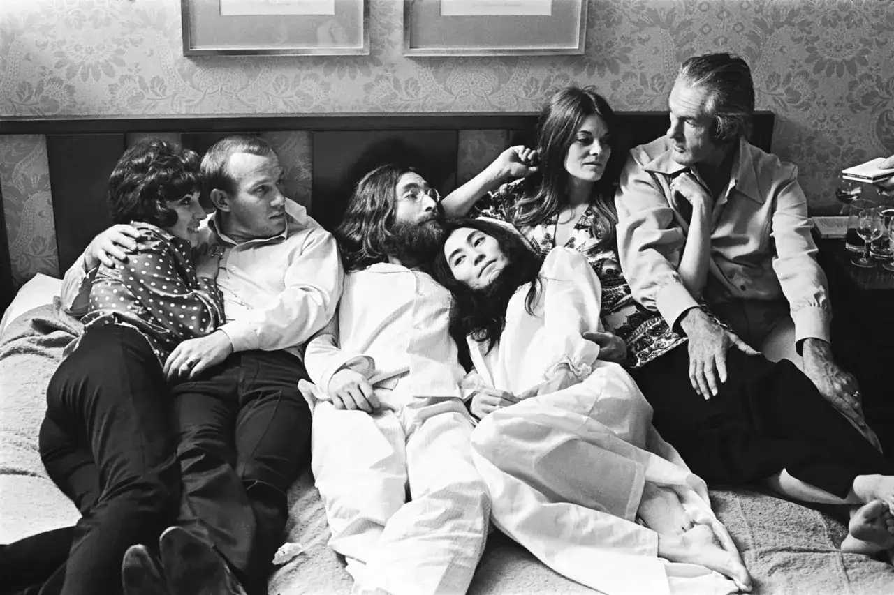 John Lennon i Yoko Ono leżą na ł&oacute;żku w otoczeniu przyjaci&oacute;ł.